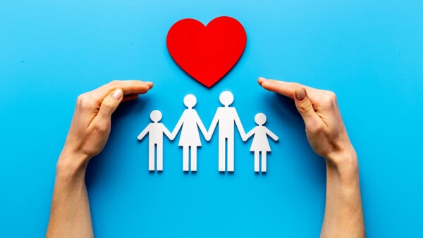 A cutout family made of paper is covered by a red paper heart and a pair of hands on top of a light blue background